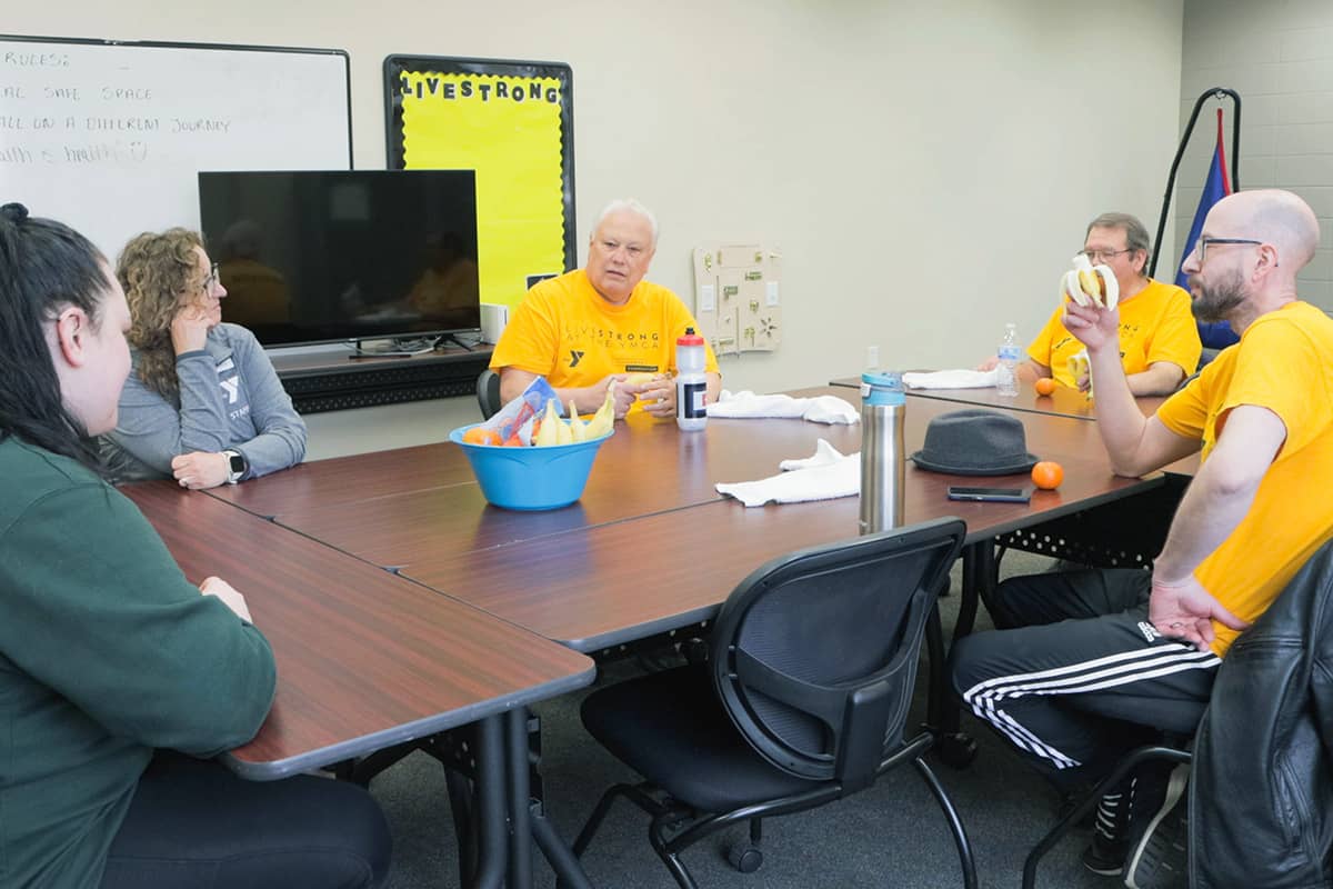 Group of adults seated around a table in a classroom setting, wearing LIVESTRONG shirts, engaged in discussion with snacks on the table.