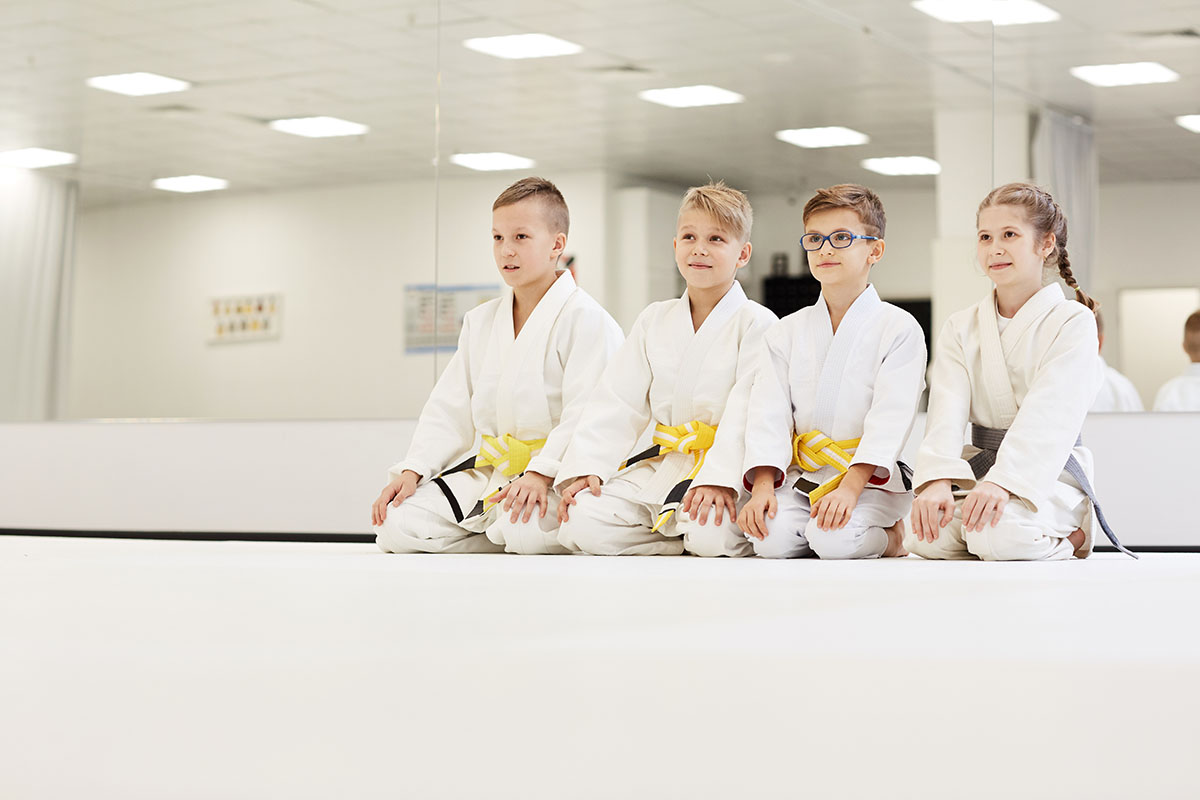 Four children wearing martial arts uniforms with yellow and gray belts, seated in a row on a white mat in a bright dojo with mirrored walls.