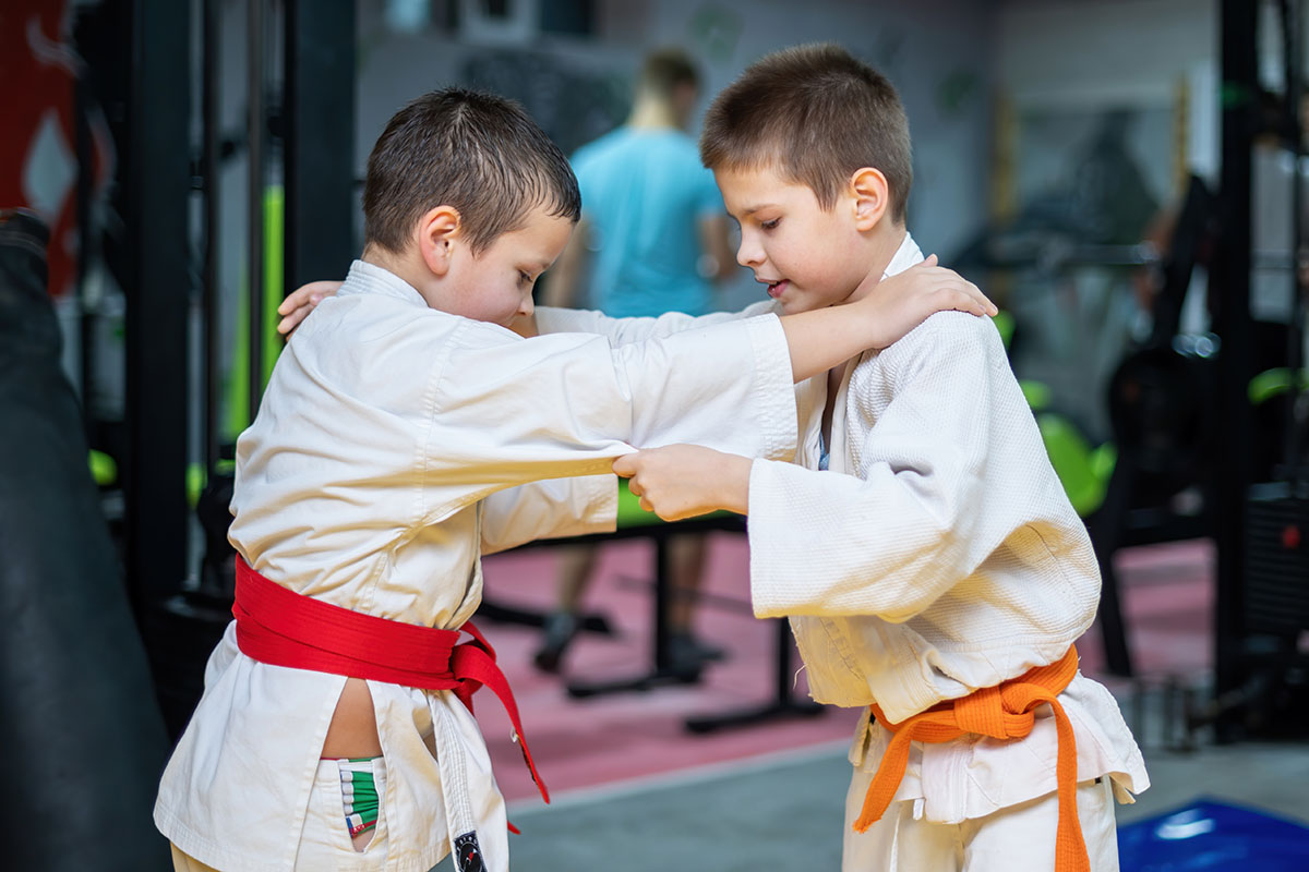 Two young boys practicing judo techniques, both wearing white gis with colored belts, in a gym environment. They appear focused and engaged in their training.