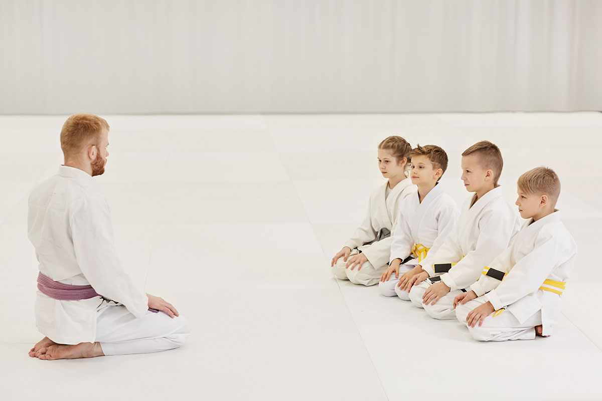 Martial arts instructor kneeling in front of a group of young students in white uniforms during a lesson.