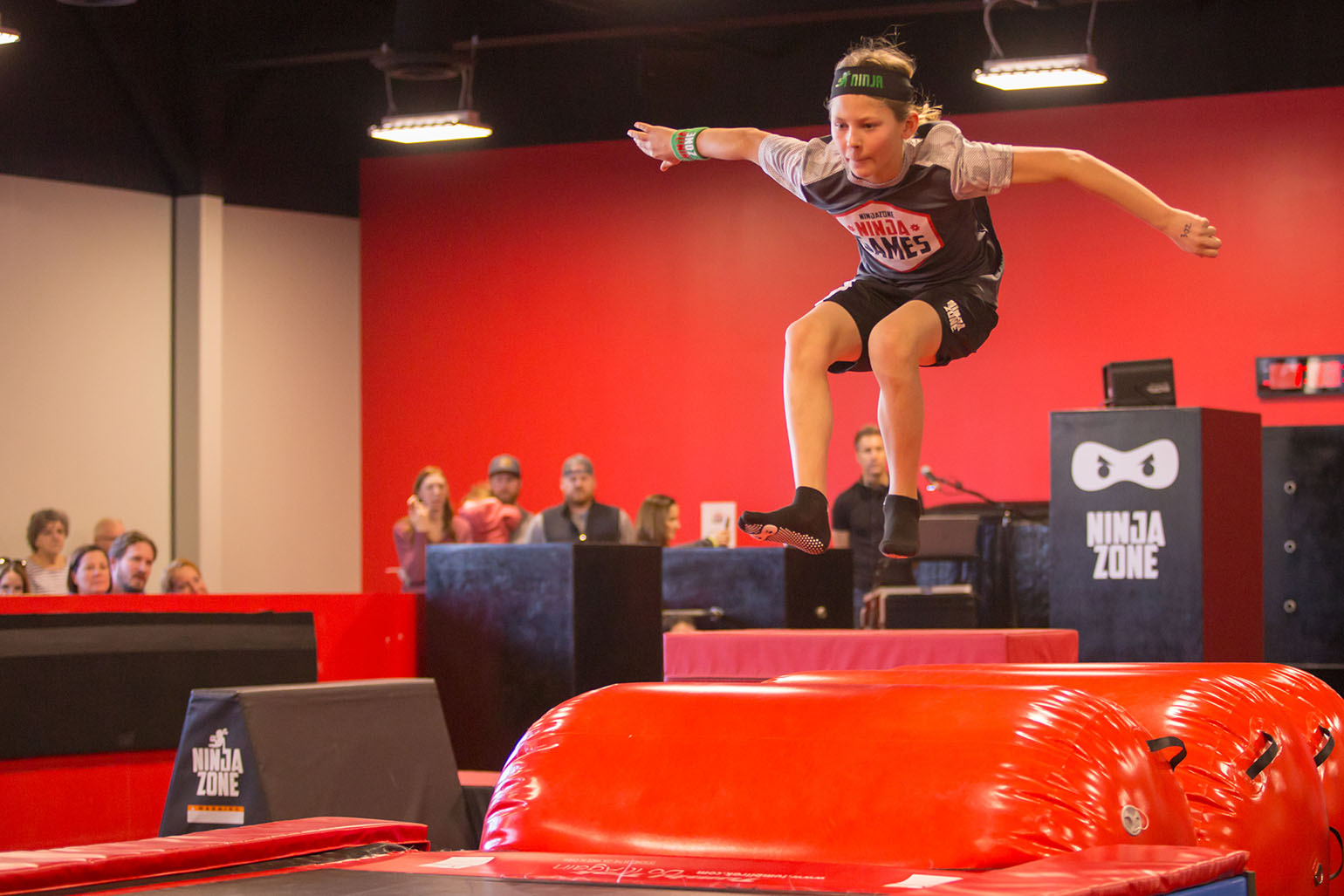 Child leaping during a Ninja Zone obstacle course event in a red and black gym.