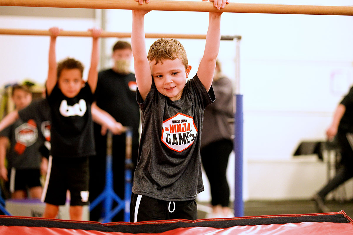 A young boy hangs from a gymnastics bar, smiling during a Ninja Games activity. Other children and instructors are visible in the background, focusing on their tasks in the training area.