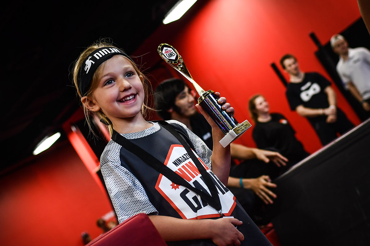 A young girl wearing a Ninja Games headband and jersey smiles while proudly holding a trophy, with a vibrant red background and other participants visible in the distance.