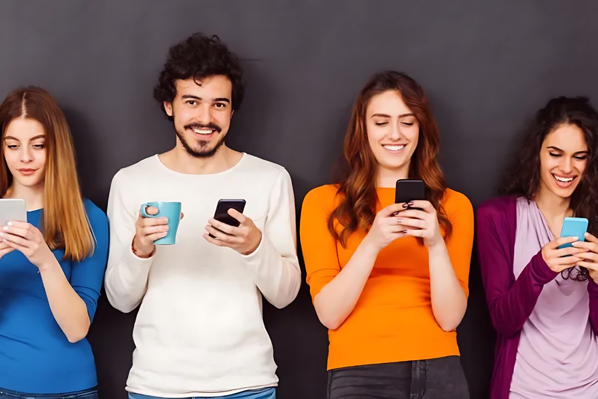 Group of four young adults standing against a dark background, smiling and using smartphones, with one holding a coffee mug.