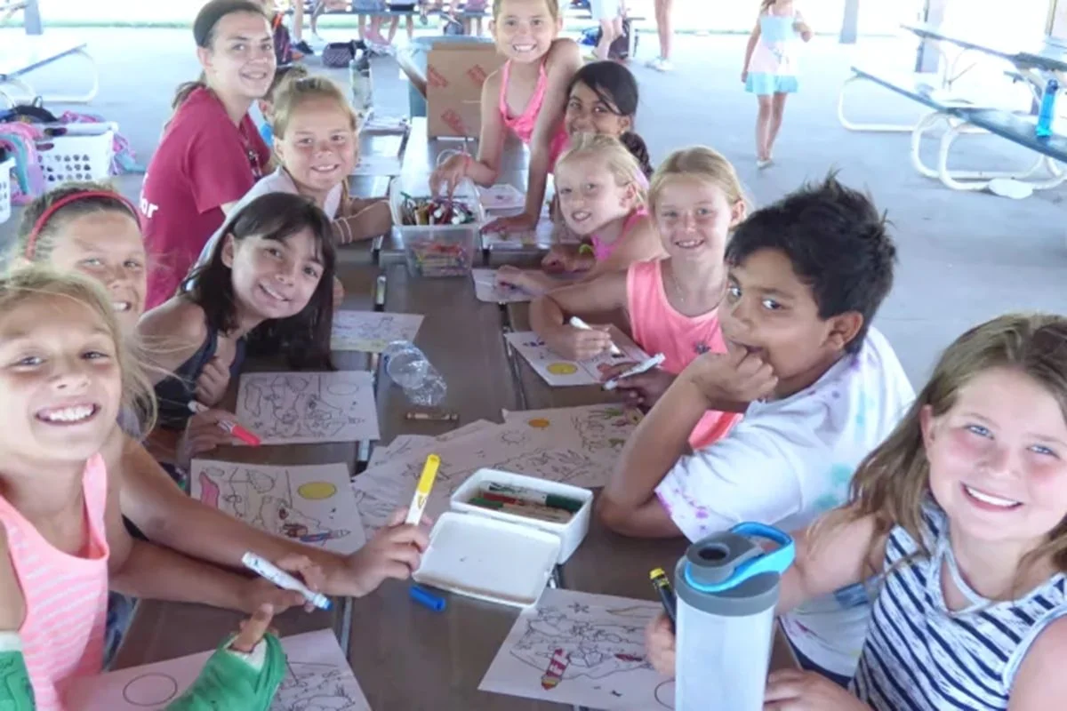 A group of children smiles while participating in a coloring activity at an outdoor table under a pavilion.