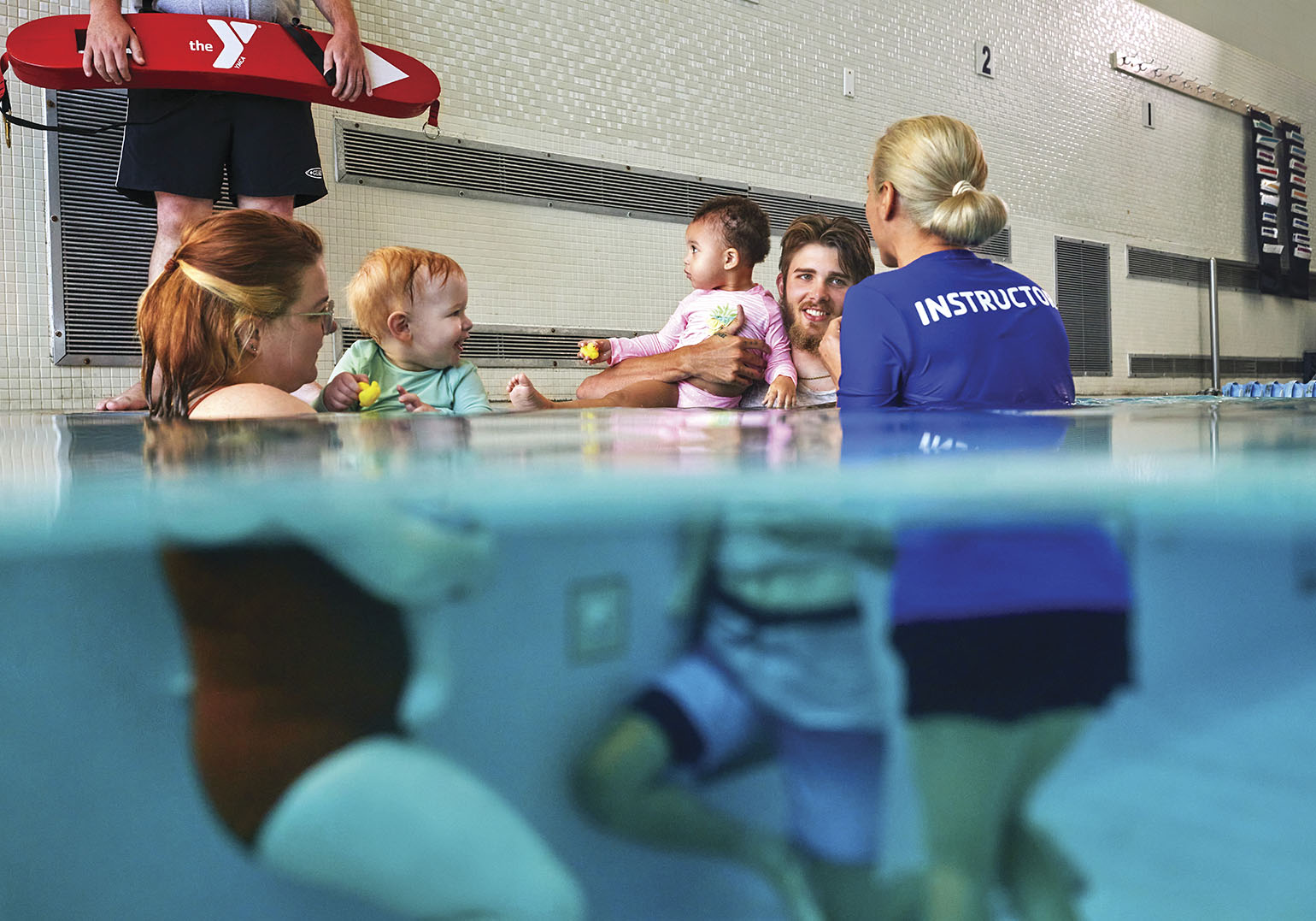 A parent-and-tot swim class at a pool, showing parents holding babies while an instructor in a blue shirt interacts with them. The photo is partially underwater, highlighting the pool's surface.