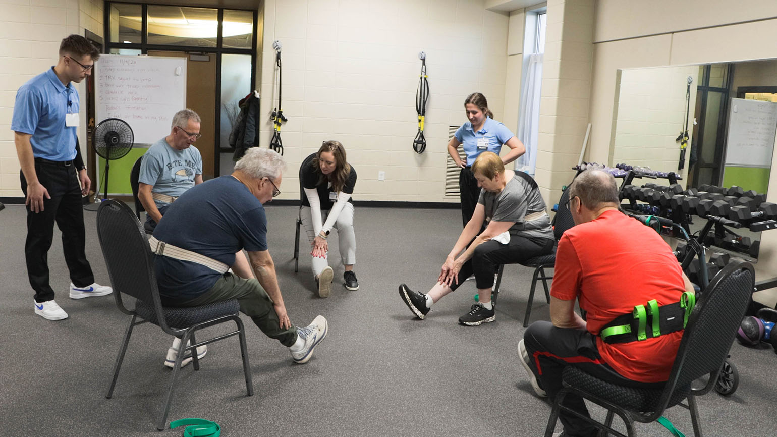 A group of older adults participates in a seated stretching exercise class, guided by two instructors in a fitness room with equipment and mirrors.