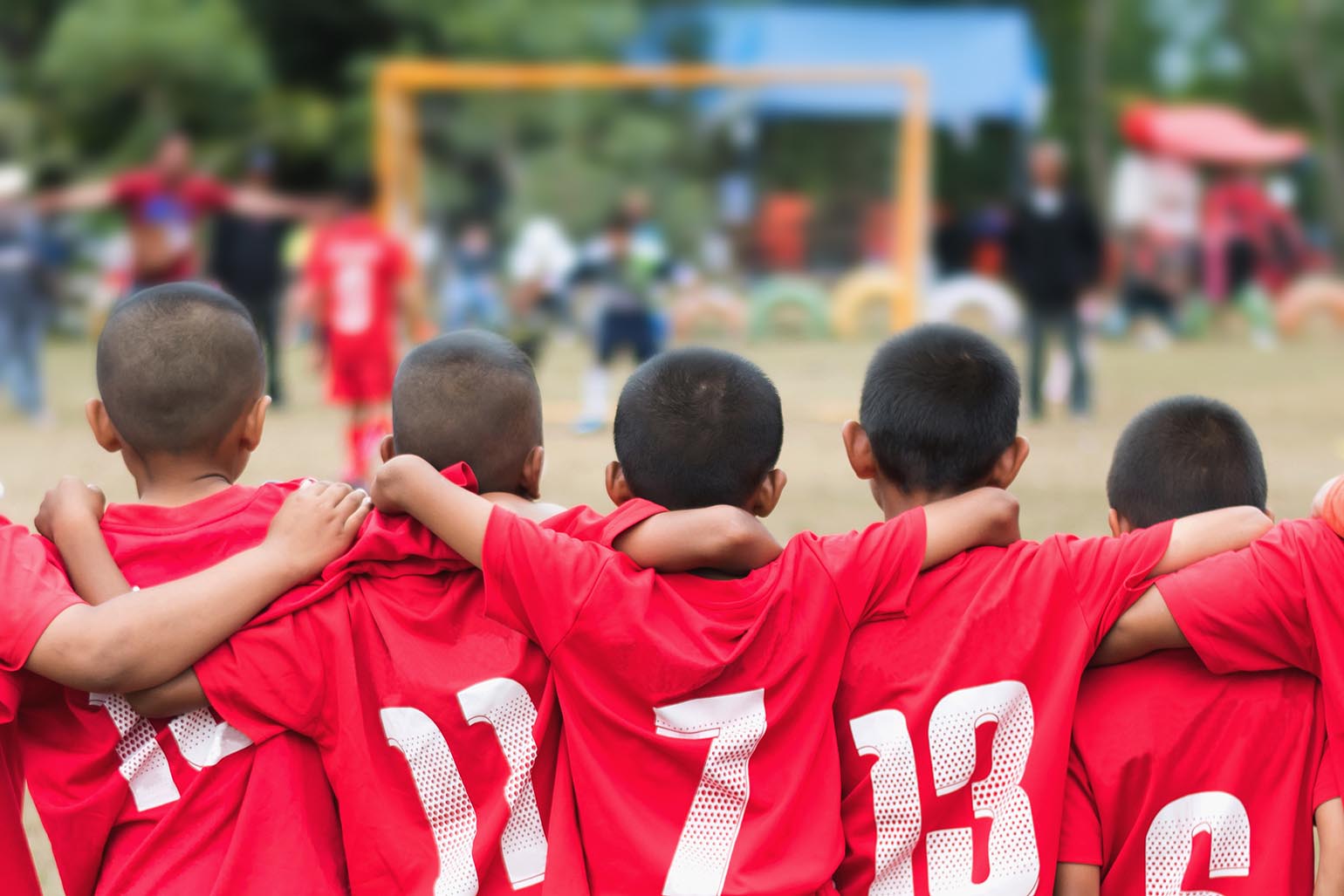 Back view of young soccer players in red jerseys with arms around each other, facing the field during a game.