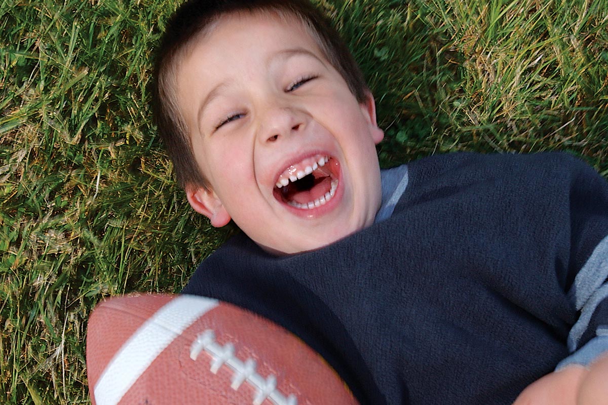 Young boy laughing on grass while holding a football.