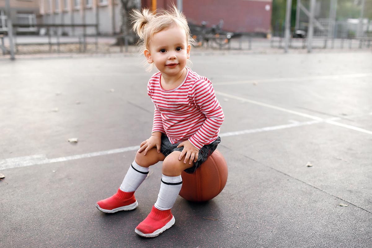 Toddler sitting on a basketball outdoors, wearing a striped shirt, shorts, and red shoes on a court.