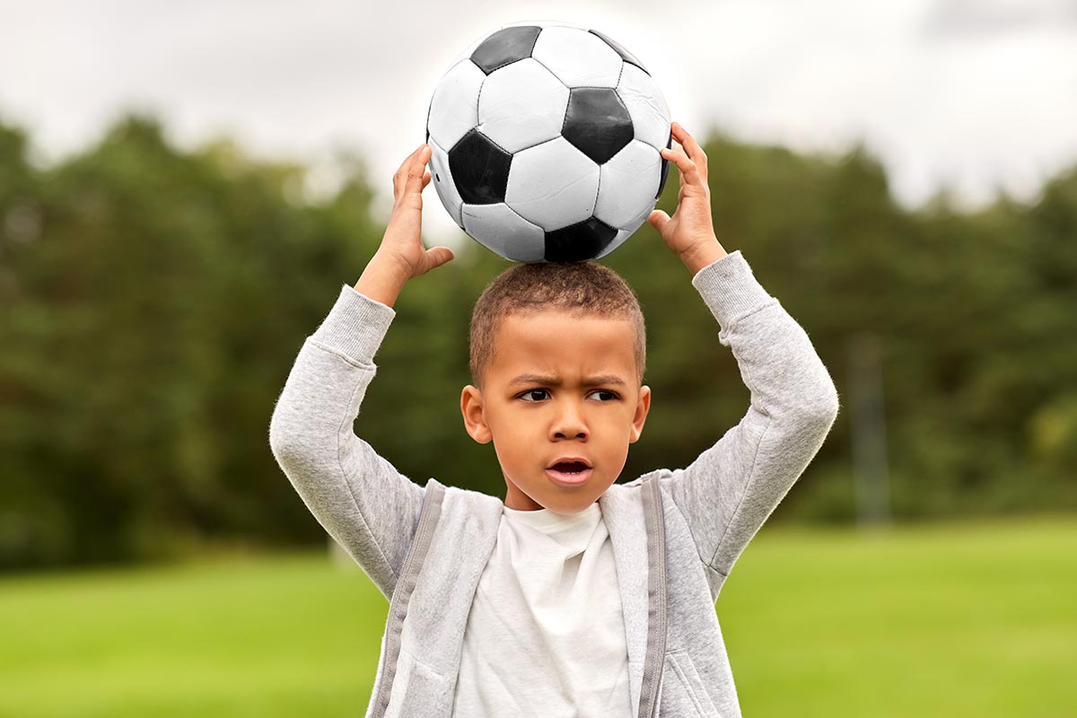 Focused young boy holding a soccer ball above his head on a grassy field.