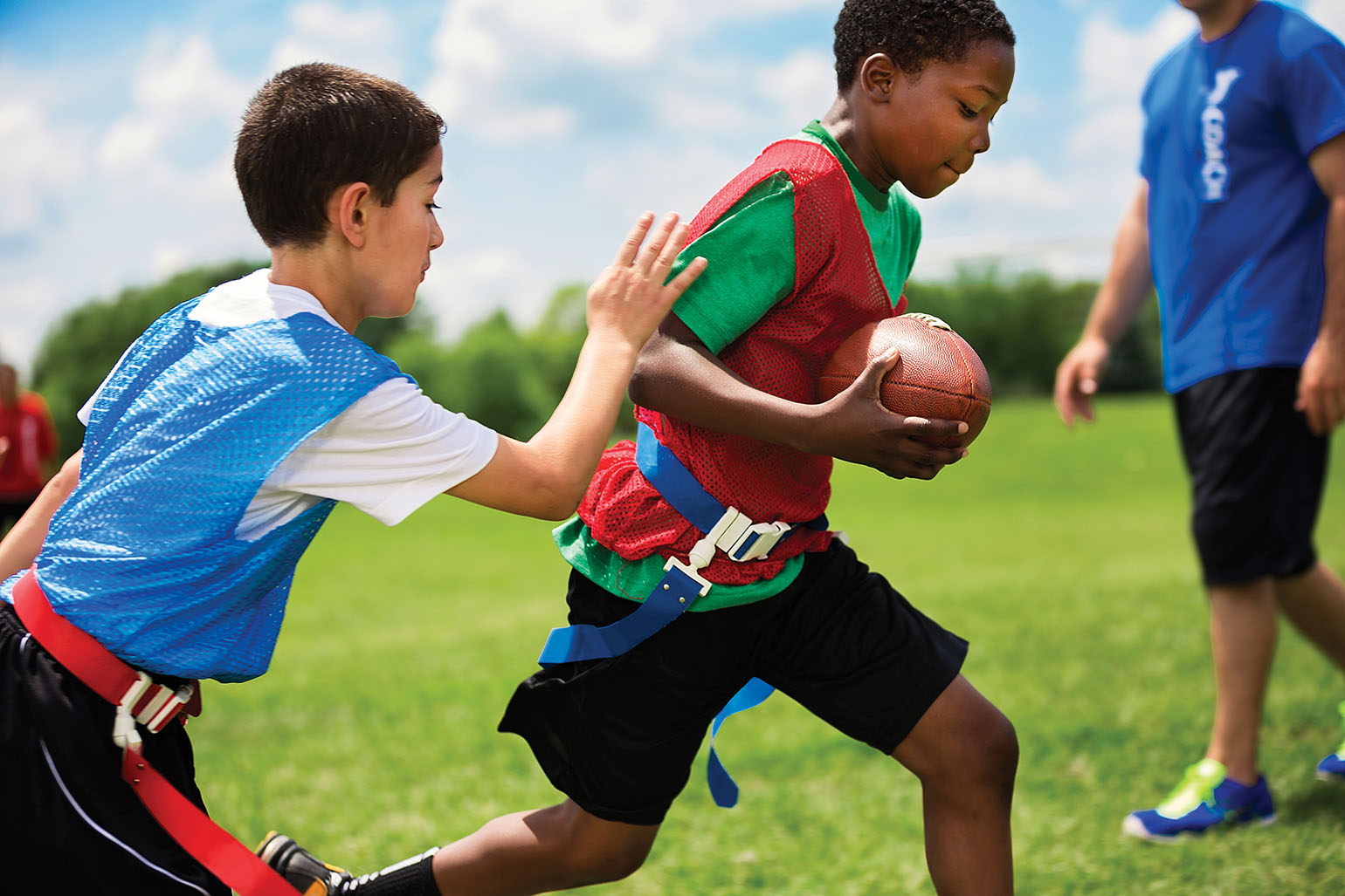 Two boys playing flag football outdoors; one running with the ball, the other reaching for the flag.