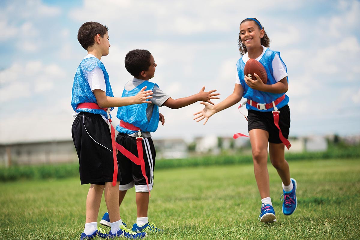 Three children playing flag football on a grassy field. One girl is running with the football and smiling, while two boys reach out to tag her. They are wearing blue practice pinnies under a bright, partly cloudy sky.