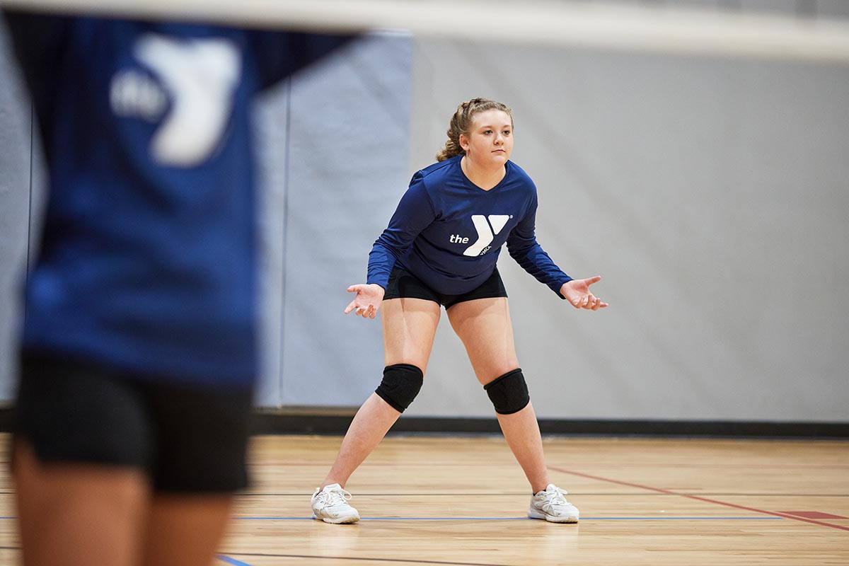 A young volleyball player wearing a blue YMCA jersey and black knee pads prepares in a ready stance on an indoor court, focused on the game, with a teammate partially visible in the foreground.