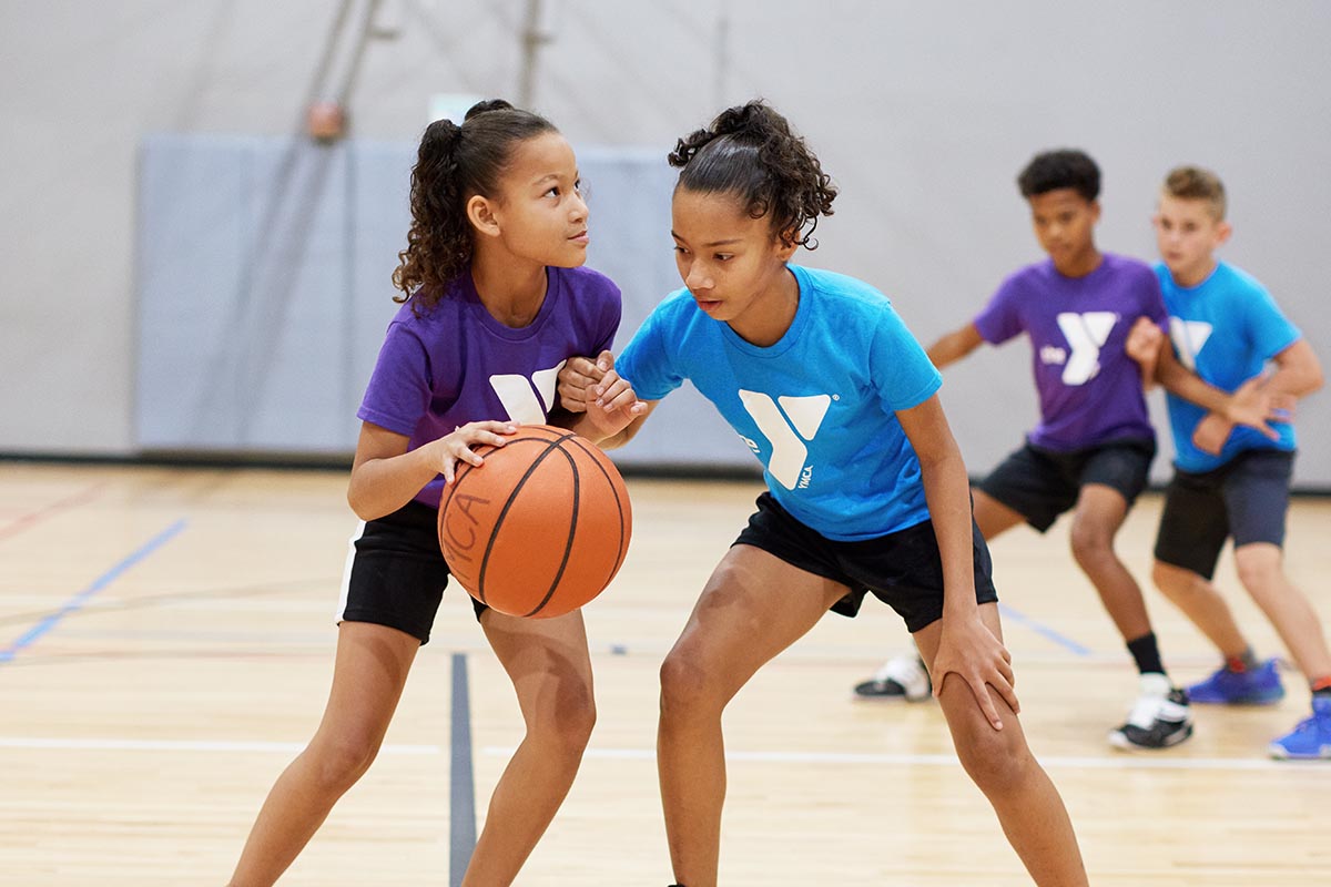 Two young girls, one wearing a purple YMCA shirt and the other a blue YMCA shirt, playing basketball in a gym. They are engaged in a defensive and offensive play, with two boys practicing in the background.