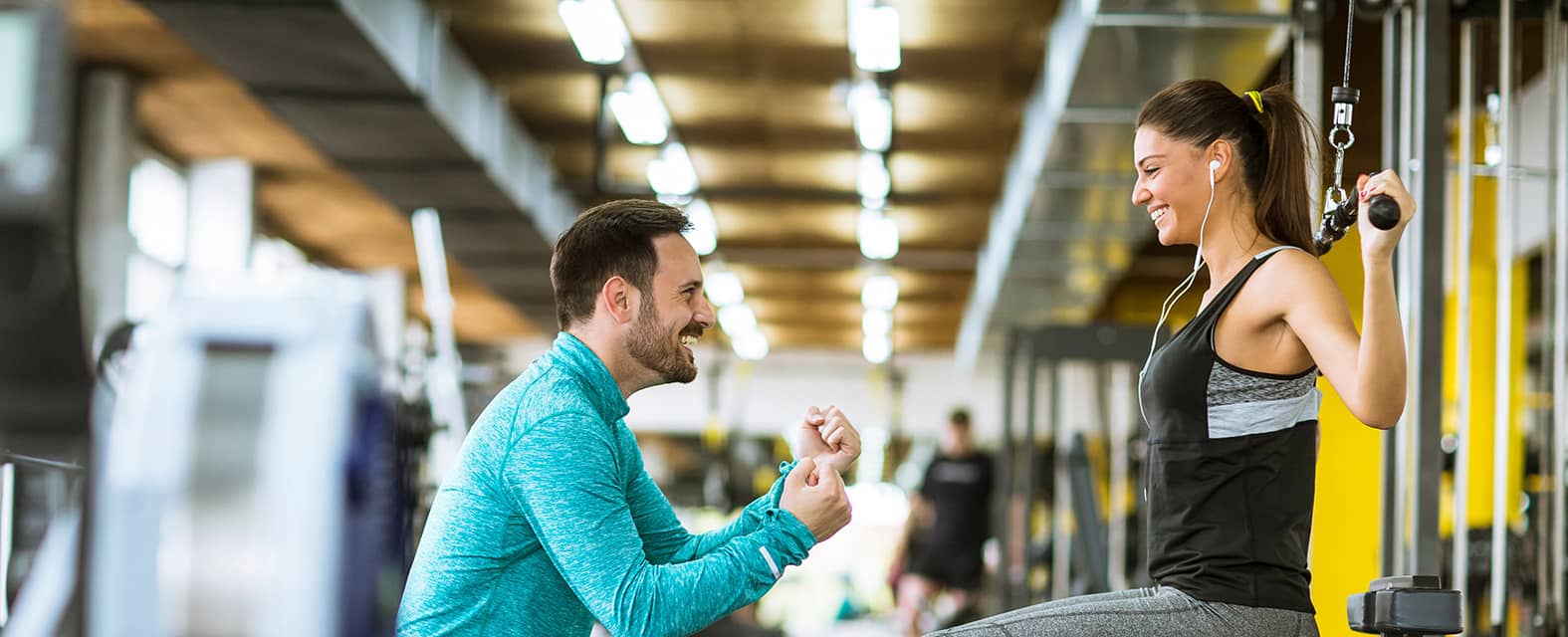 A personal trainer in a blue sweatshirt encourages a smiling woman using a cable machine at the gym.