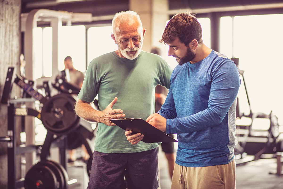 A fitness trainer discusses a workout plan with an older man in a gym, surrounded by exercise equipment.