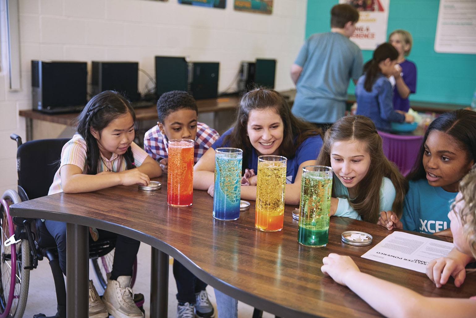 A group of children and an instructor gathered around a table, observing colorful science experiments in tall glass containers. A child in a wheelchair is part of the group, creating an inclusive learning environment. Computers and posters decorate the classroom in the background.