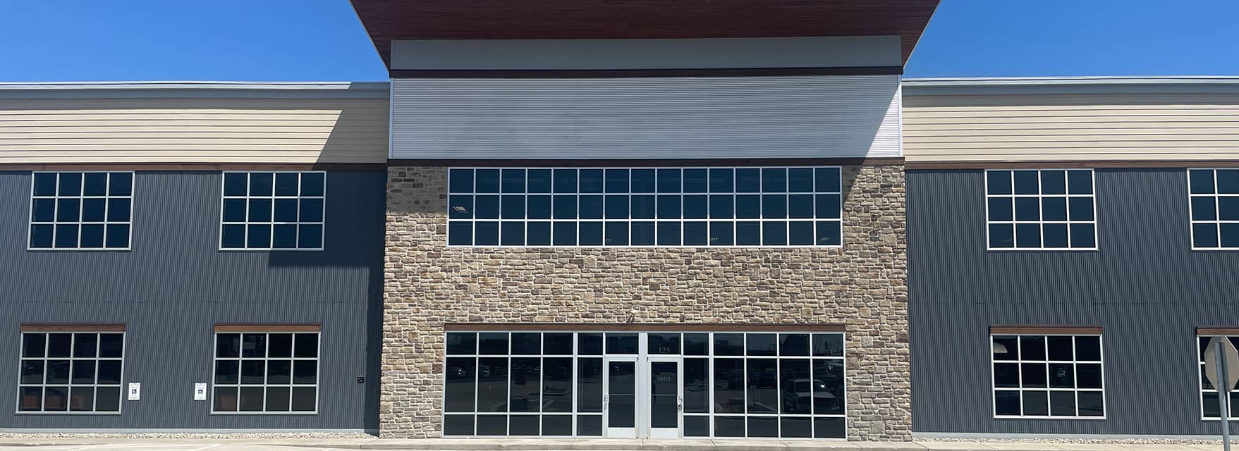 Front view of a modern building with large windows, stone accents, and a metal facade under a clear blue sky.