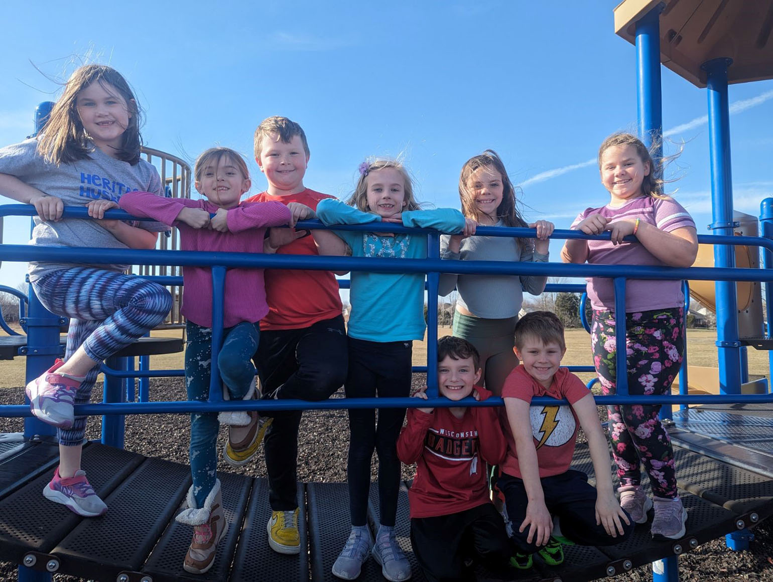 A group of smiling children poses together on a playground structure under a clear blue sky.