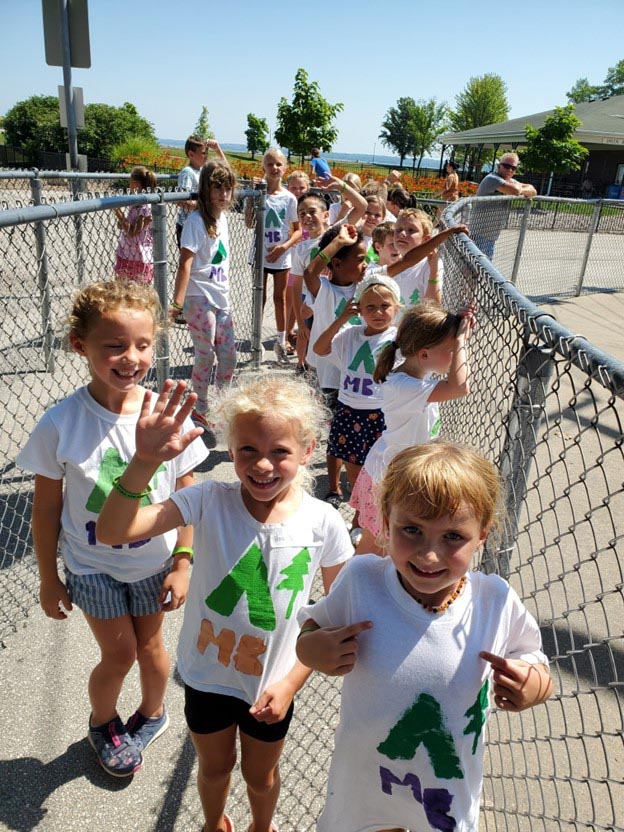 Group of children wearing matching camp shirts, smiling and waving while standing in a fenced outdoor area on a sunny day.