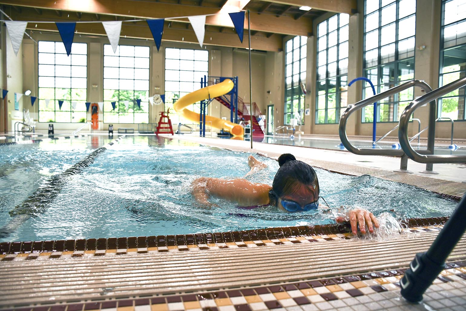 A swimmer wearing goggles practices freestyle strokes in an indoor pool with a water slide and large windows in the background.