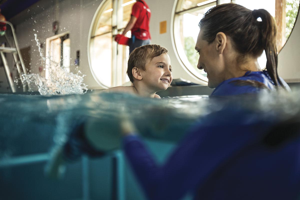 A smiling child and an adult share a moment during a swimming lesson in an indoor pool, with a lifeguard in the background.