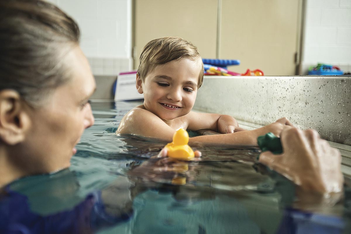 Young boy smiling in a pool during a swim lesson, playing with a yellow rubber duck held by his instructor.