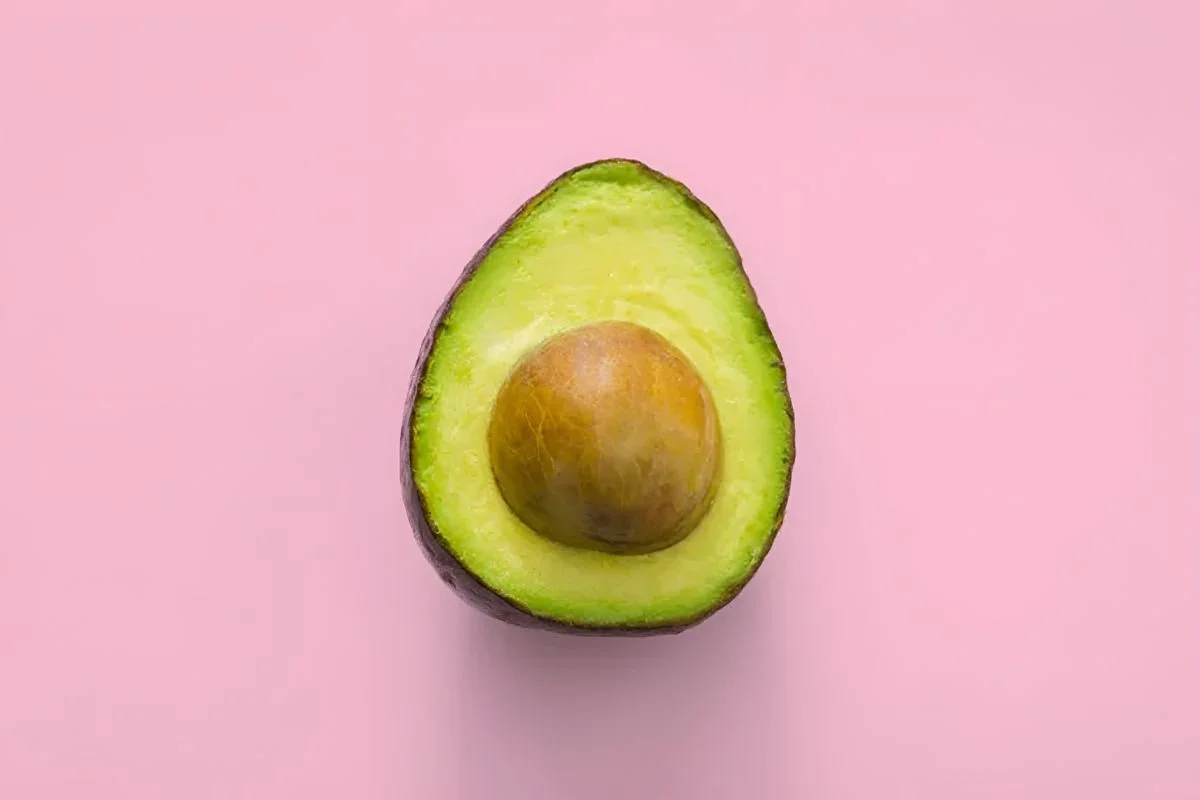 A halved avocado with a visible pit, placed against a soft pink background.