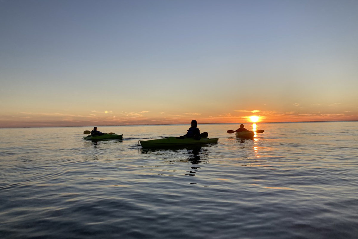 Three people kayak on calm water at sunset, silhouetted against the glowing horizon.