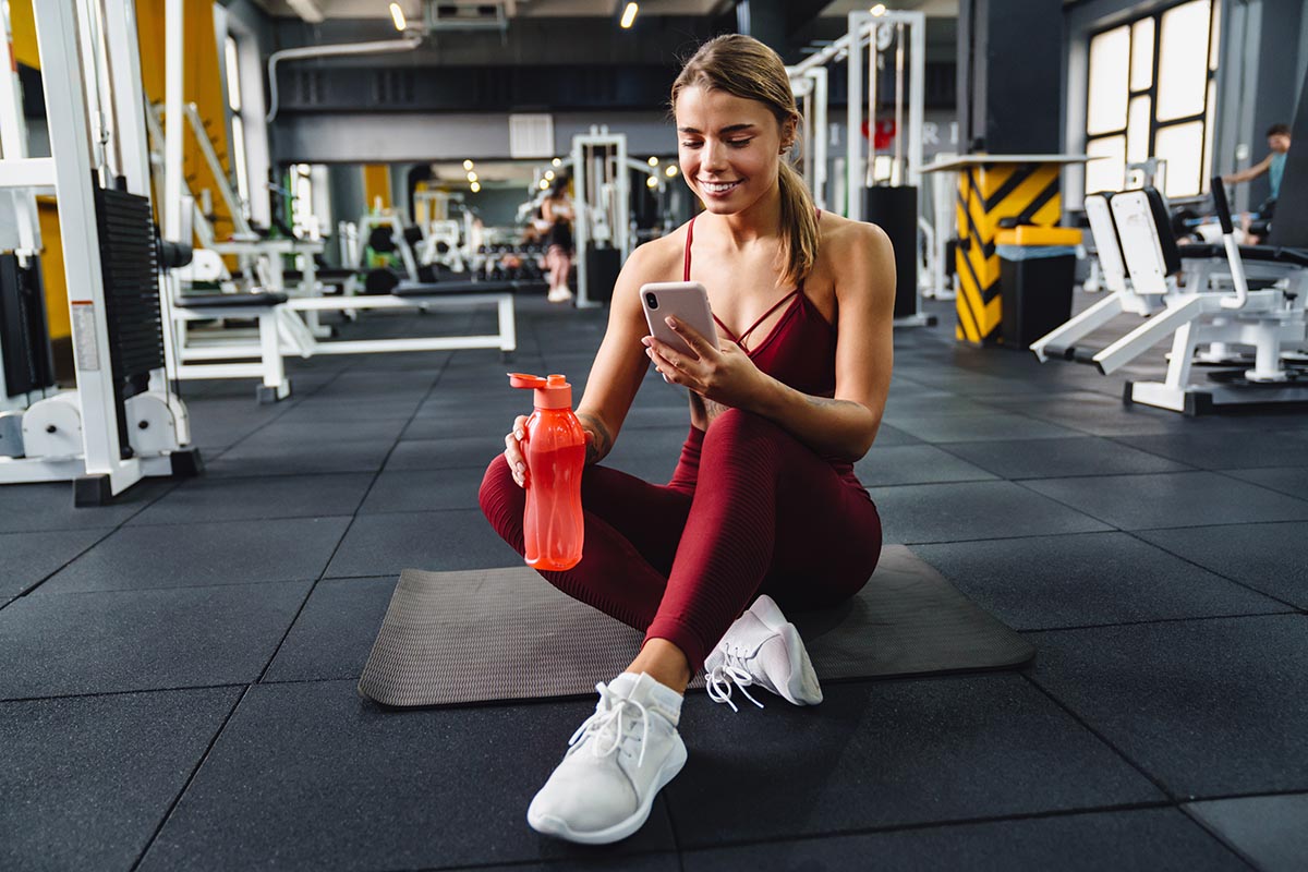 A woman in workout attire sits on a yoga mat in a gym, holding a water bottle and looking at her phone. Exercise equipment surrounds her in the modern fitness space.