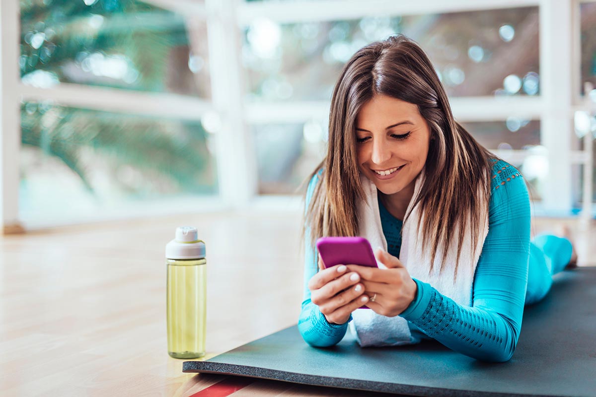 Woman lying on a yoga mat in a bright gym, smiling while using a smartphone, with a water bottle nearby.