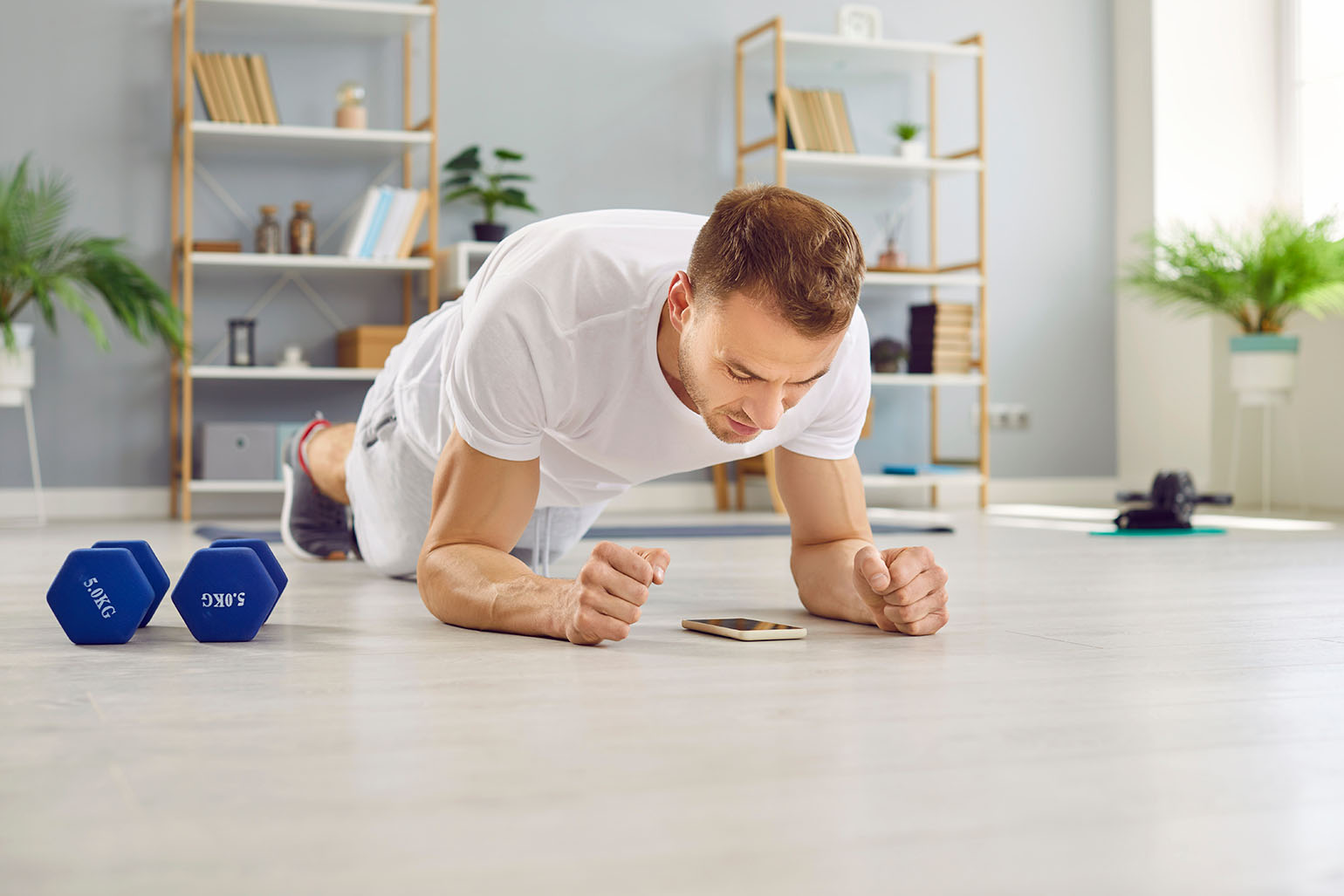 Young man using timer or fitness workout app on mobile phone while doing low forearm elbow plank position exercise for abdominal muscles on floor in living room with dumbbells and other equipment