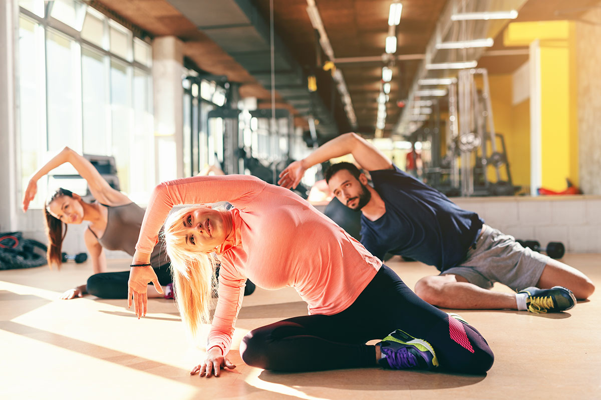 A group of people, including a pregnant woman in the foreground, stretch during a fitness class in a bright, spacious gym with large windows and modern equipment.