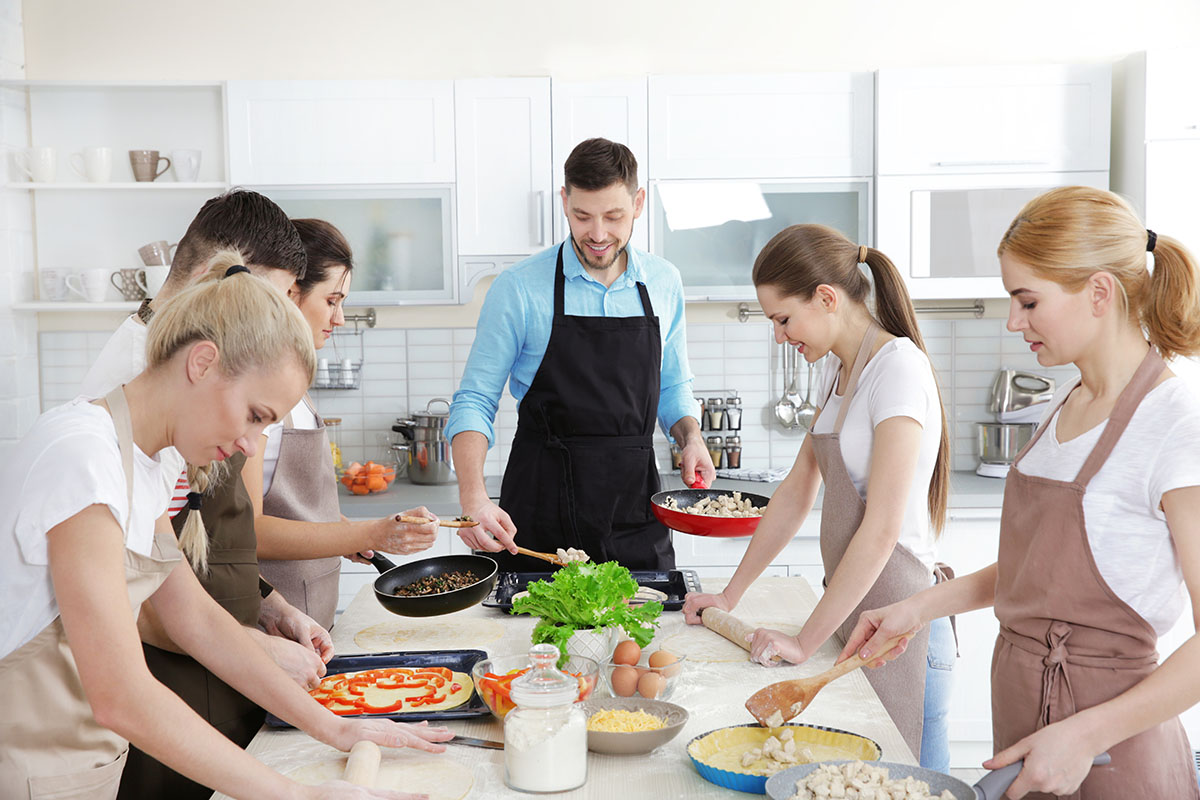 A group of adults in a bright kitchen participating in a cooking class. They wear aprons and work together to prepare various dishes, including pizza and sautéed vegetables. Ingredients like eggs, lettuce, and flour are spread across the table.