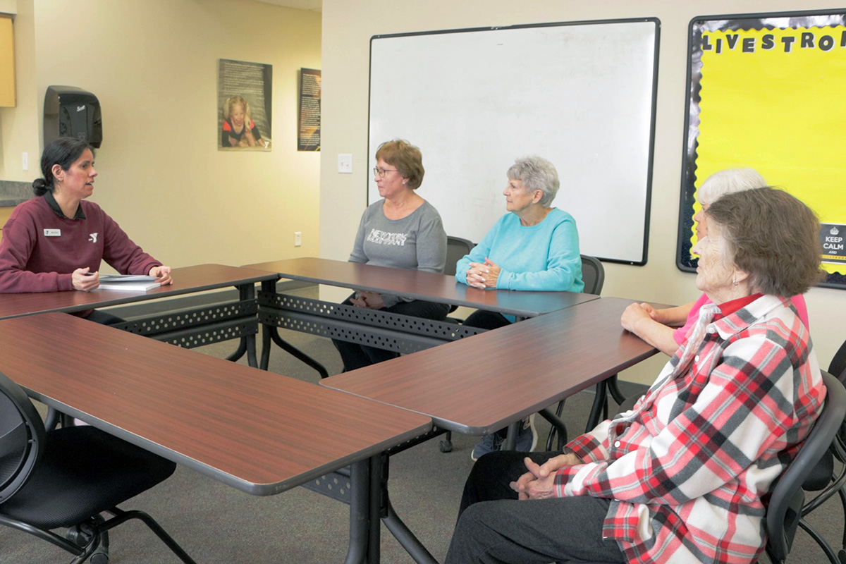 A facilitator leads a small discussion group of older adults seated around a U-shaped table in a classroom setting.