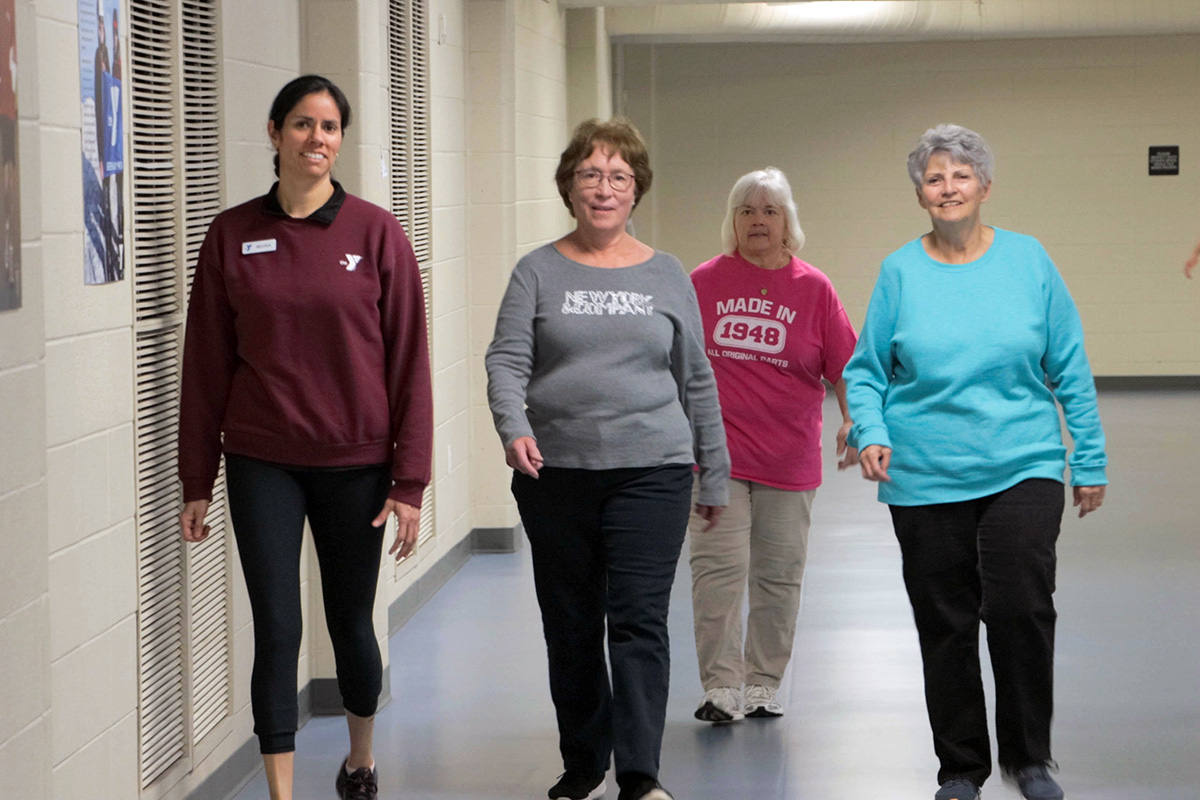 Group of women walking indoors for fitness, led by a smiling instructor wearing YMCA apparel.