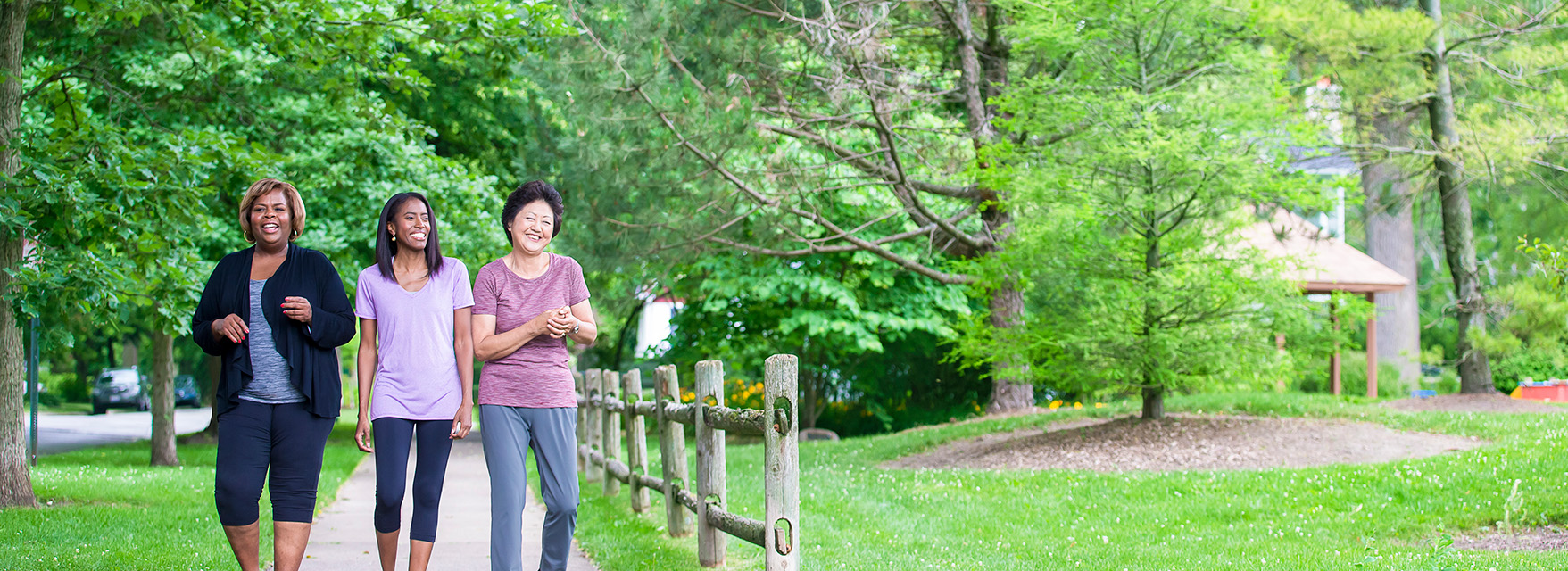 Three women enjoy a walk on a shaded path in a green park, surrounded by trees and a wooden fence.