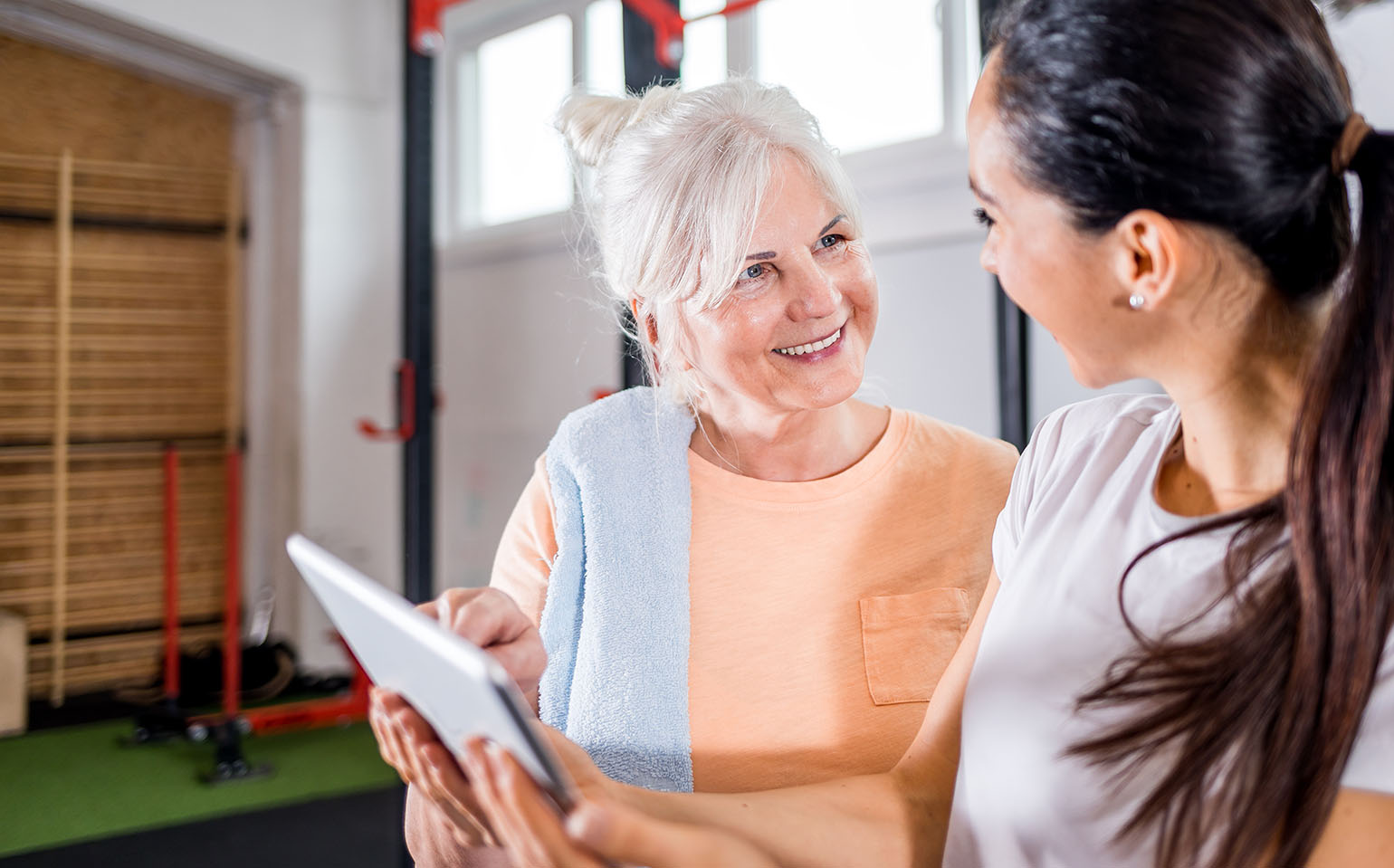 A senior woman with a towel over her shoulder smiles as she discusses a workout plan with a personal trainer in a gym setting.