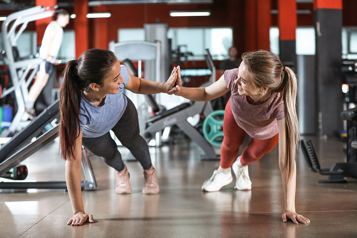 Two women doing partner push-ups and high-fiving in a gym environment.