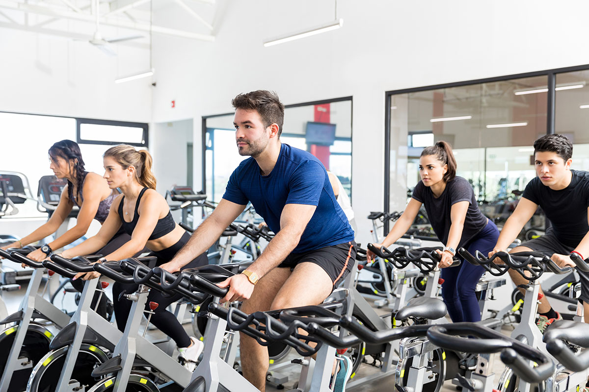 Group of people focused on a spin class, riding stationary bikes in a bright, modern gym with large windows and mirrors.