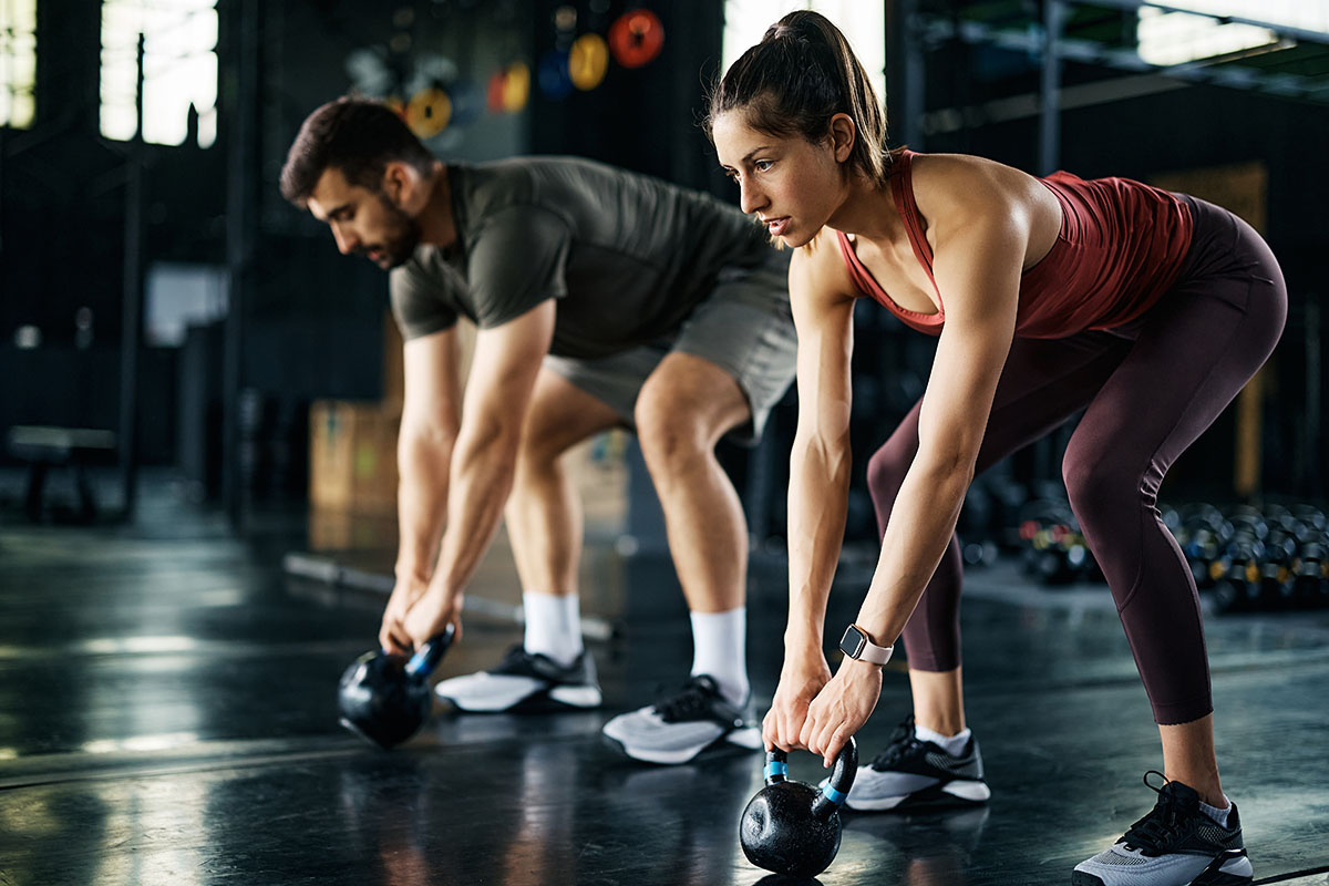 A man and a woman performing kettlebell exercises in a gym. Both are focused on their workout, bent over with kettlebells in hand, in a well-lit, modern fitness environment.