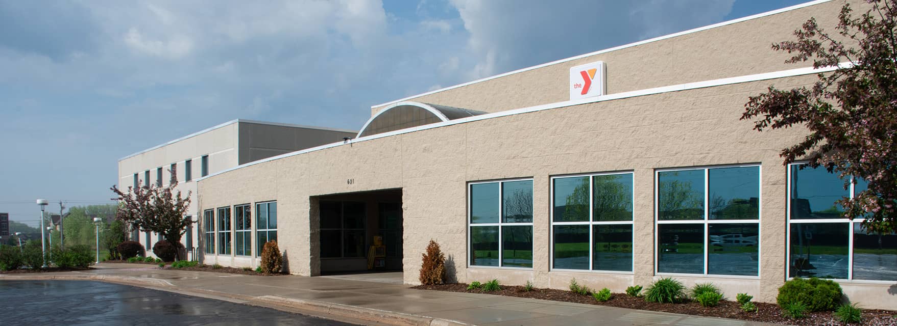 The exterior of a YMCA building with large windows, surrounded by landscaping and a paved entrance under a partly cloudy sky.