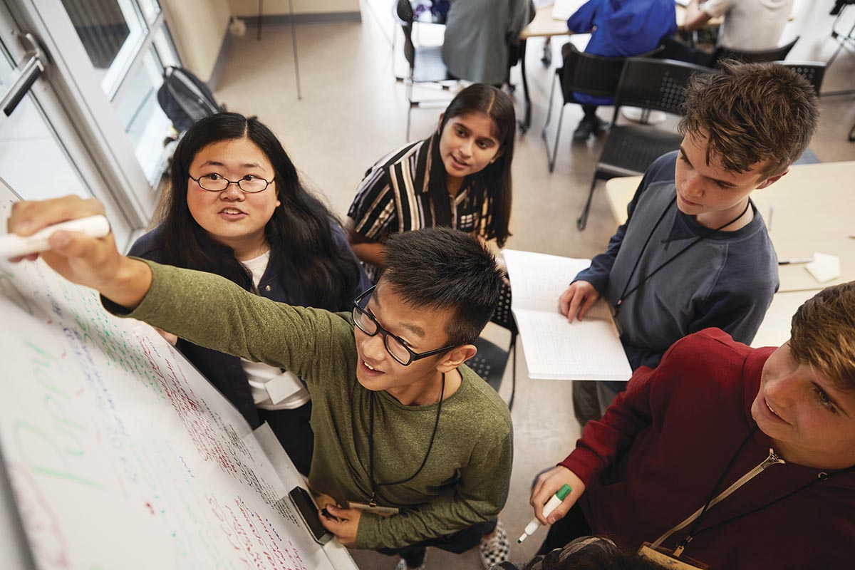 A group of students collaborates on a project, writing and brainstorming ideas on a whiteboard in a classroom setting.