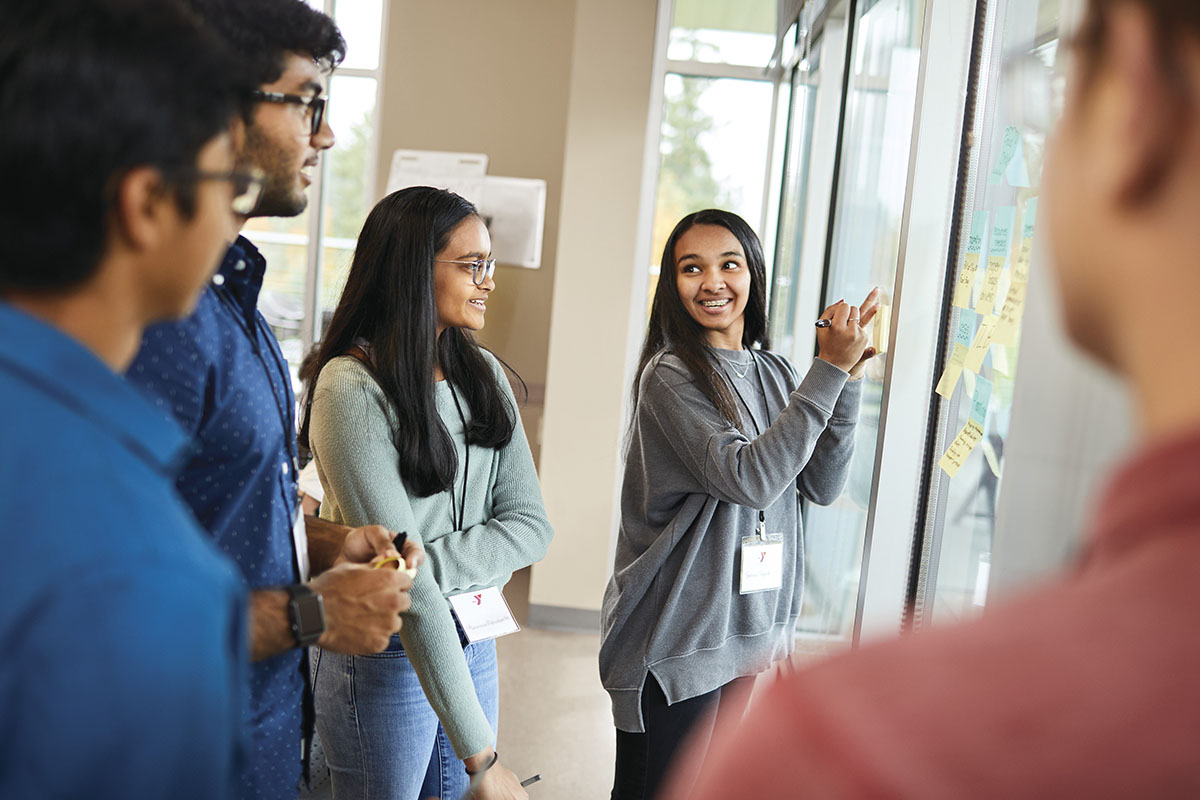 Group of young adults collaborating near a glass wall with sticky notes, one person writing while others listen and engage.