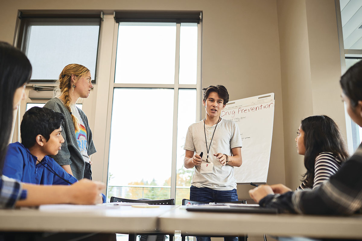 Young facilitator leading a group discussion on drug prevention at a YMCA program, with participants seated around a table and a presentation board in the background.