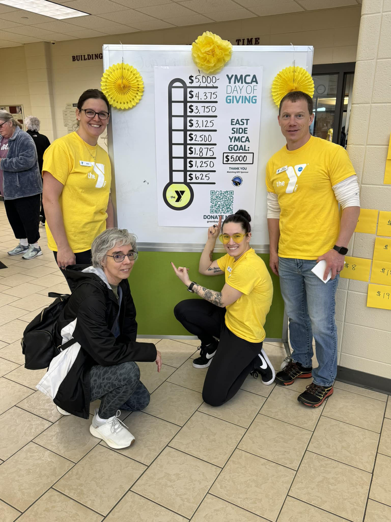 Four individuals posing near a donation progress board for YMCA Day of Giving, celebrating fundraising efforts in a community center.