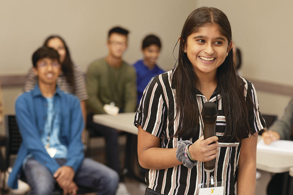 Teen girl speaking into a microphone with peers seated behind her in a classroom setting.