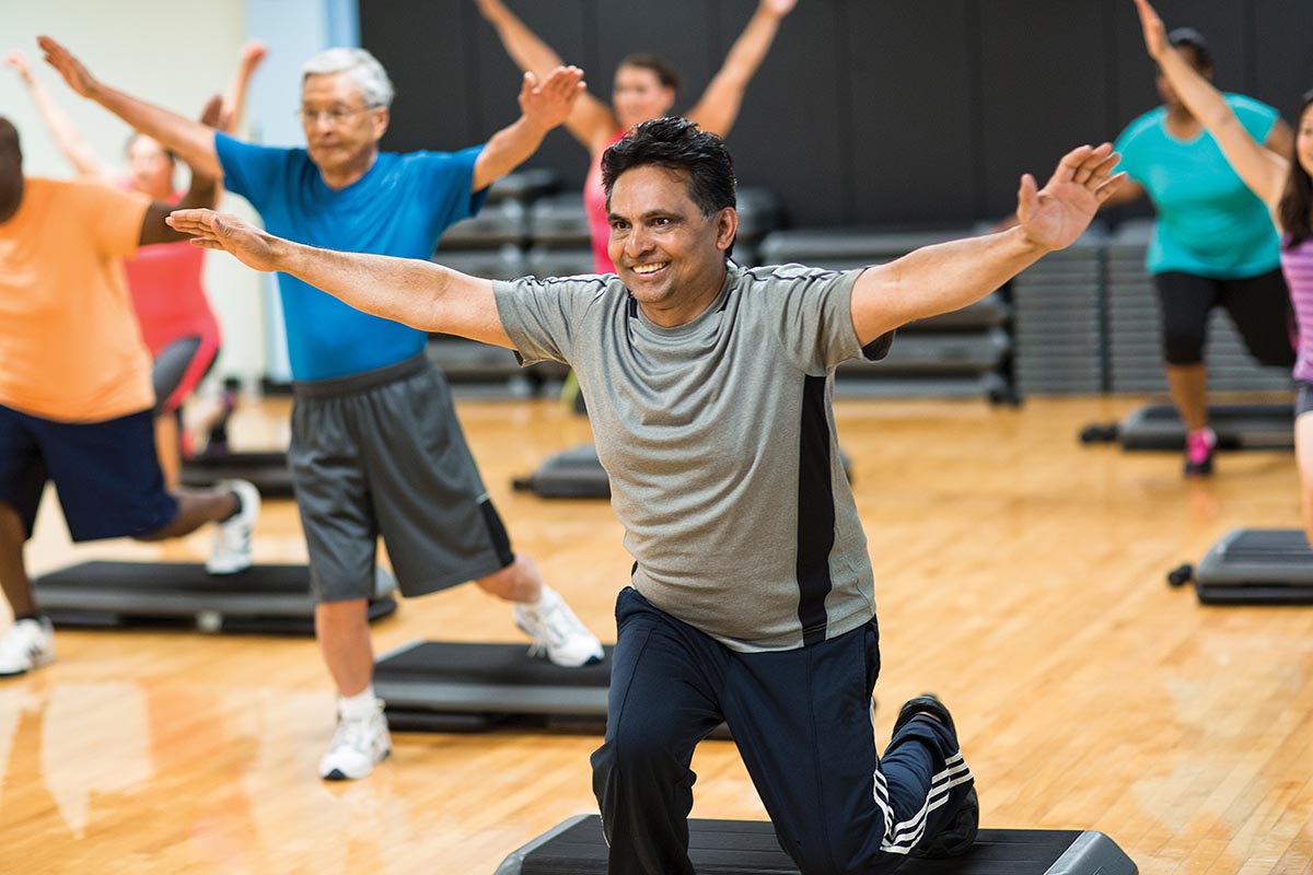 Group fitness class participants smiling while balancing on one leg with arms extended in a gym setting.