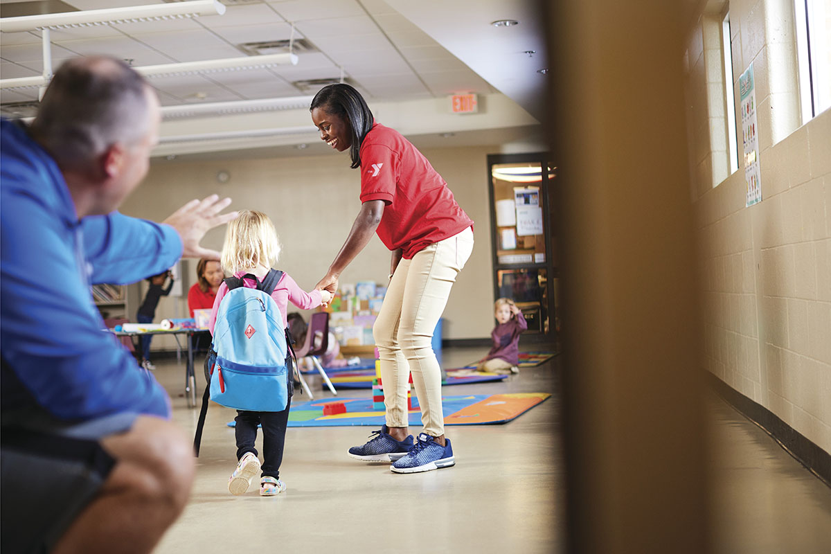 A young child with a blue backpack being guided into a classroom by a smiling staff member in a red shirt, while an adult waves goodbye in the foreground.